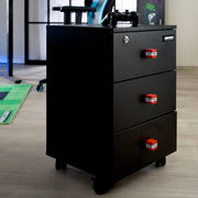 Schwarzer Rollcontainer mit drei Schubladen in Carbon-Optik und roten TNT-Griffen, mit Schloss an der oberen Schublade; Perspektive: frontale Dreiviertelansicht von links auf Bodenhöhe.