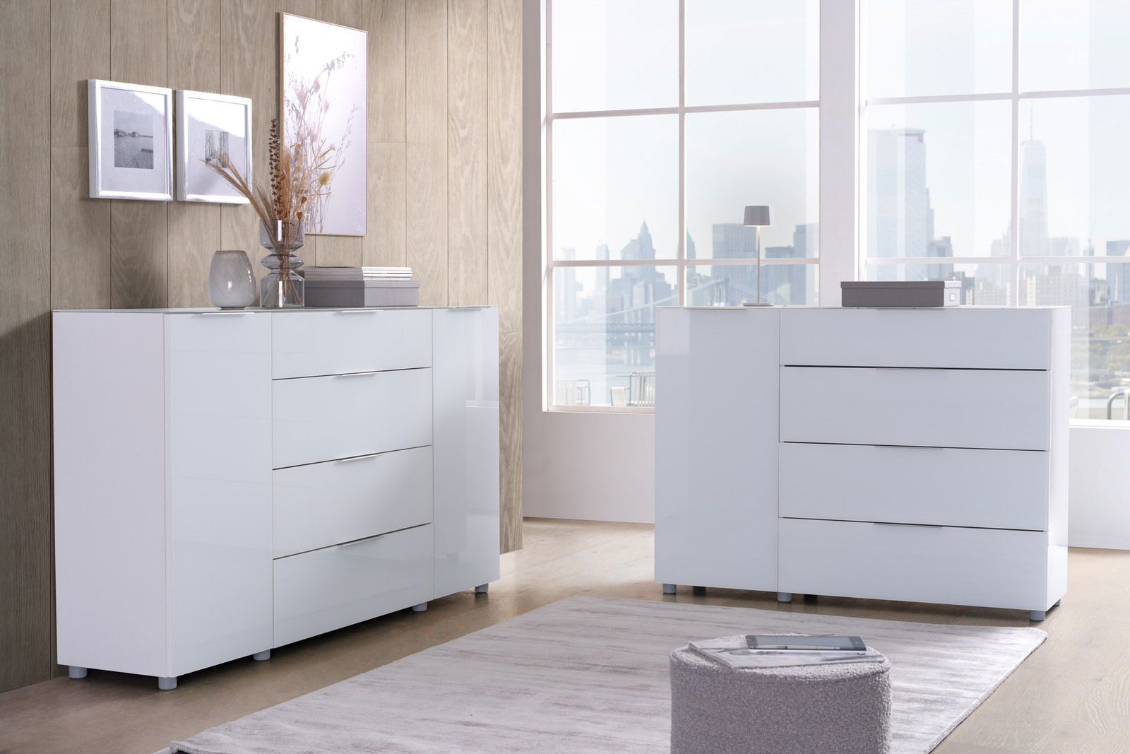 Voleo Sideboard MALIN Zwei moderne, weiße Kommoden in einem hellen Raum mit großen Fenstern, die eine Stadtansicht bieten. Die Kommoden haben eine glänzende Oberfläche und sind aus der Perspektive leicht seitlich zu sehen.