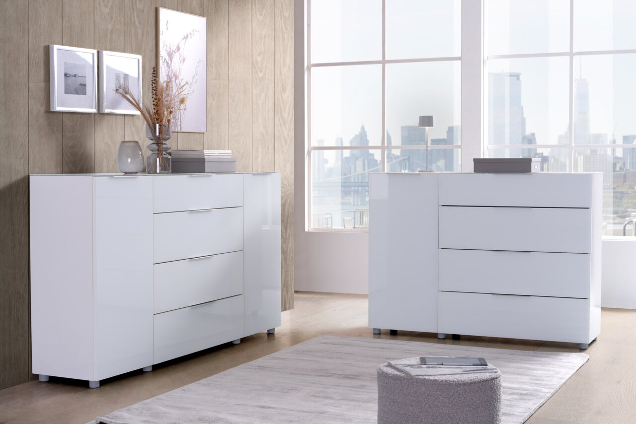 Voleo Sideboard MALIN Zwei moderne, weiße Kommoden in einem hellen Raum mit großen Fenstern, die eine Stadtansicht bieten. Die Kommoden haben eine glänzende Oberfläche und sind aus der Perspektive leicht seitlich zu sehen.