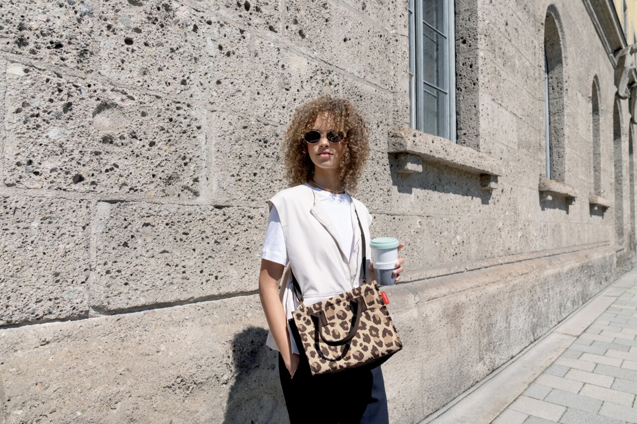 Reisenthel Daily Shopper XS Leo Macchiato Frau mit lockigem Haar und Sonnenbrille trägt eine kleine Leopardenmuster-Tasche über der Schulter und hält einen Kaffeebecher, während sie an einer Steinmauer entlanggeht.