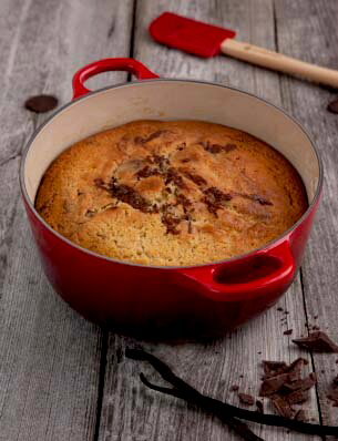 Roter runder Le Creuset Bräter, 24 cm, mit gebackenem Kuchen, von oben fotografiert, auf rustikalem Holztisch mit Schokoladenstücken und Vanilleschoten.