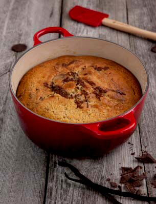 Roter runder Le Creuset Bräter, 24 cm, mit gebackenem Kuchen, von oben fotografiert, auf rustikalem Holztisch mit Schokoladenstücken und Vanilleschoten.