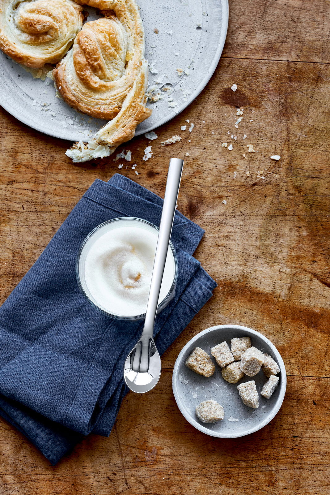 WMF Latte Macchiato-Löffel 6-tlg. NUOVA Latte Macchiato-Löffel Nuova auf einem Holztisch, neben einem Glas mit Milchschaum und Gebäck, aus der Vogelperspektive.