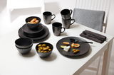 Schwarzes Geschirrset auf einem weißen Tisch, bestehend aus Desserttellern, Schalen und Tassen, mit Oliven und Früchten serviert, aus seitlicher Perspektive fotografiert.