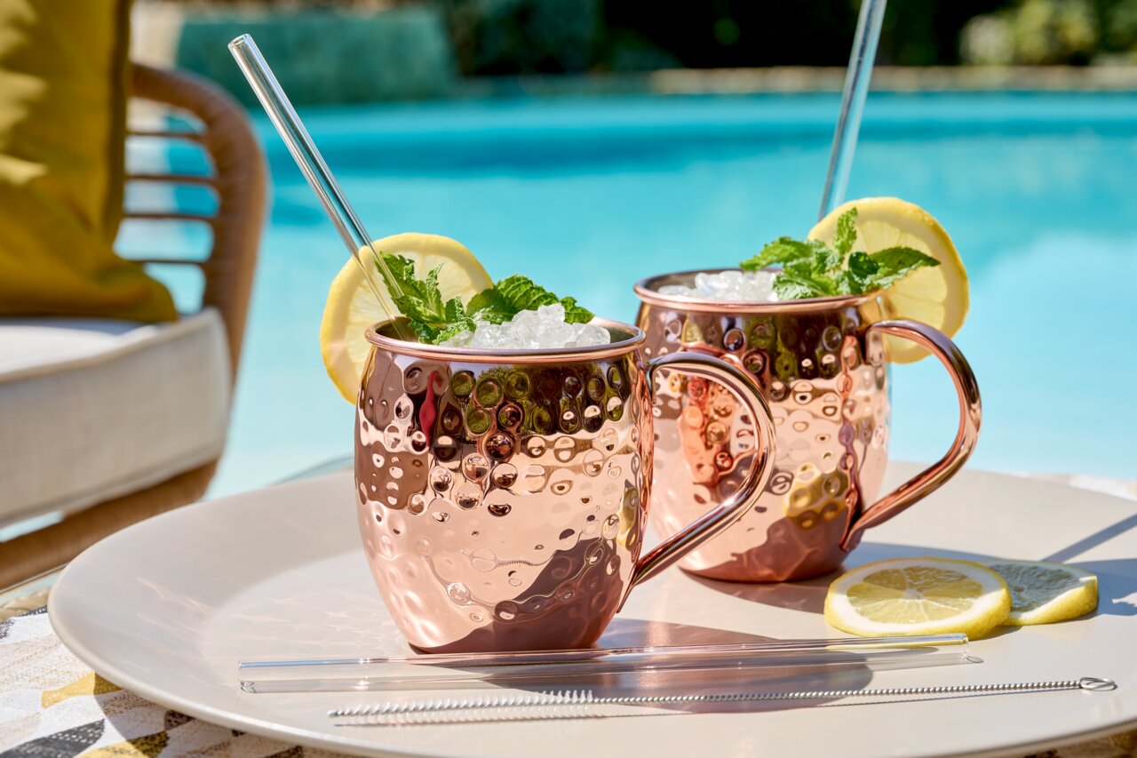 Zwei gehämmerte Kupferbecher mit Griff, gefüllt mit Eis, Minze und Zitronenscheiben, auf einem Tablett am Pool, seitliche Perspektive.