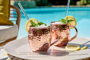 Zwei gehämmerte Kupferbecher mit Griff, gefüllt mit Eis, Minze und Zitronenscheiben, auf einem Tablett am Pool, seitliche Perspektive.