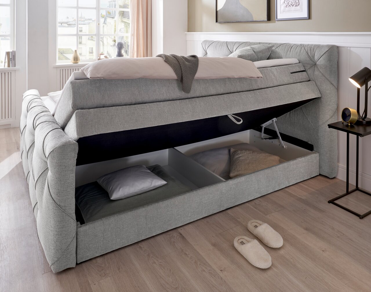 Boxspringbett mit geöffnetem Stauraum, seitliche Perspektive, grauer Stoffbezug, Tageslicht durch Fenster