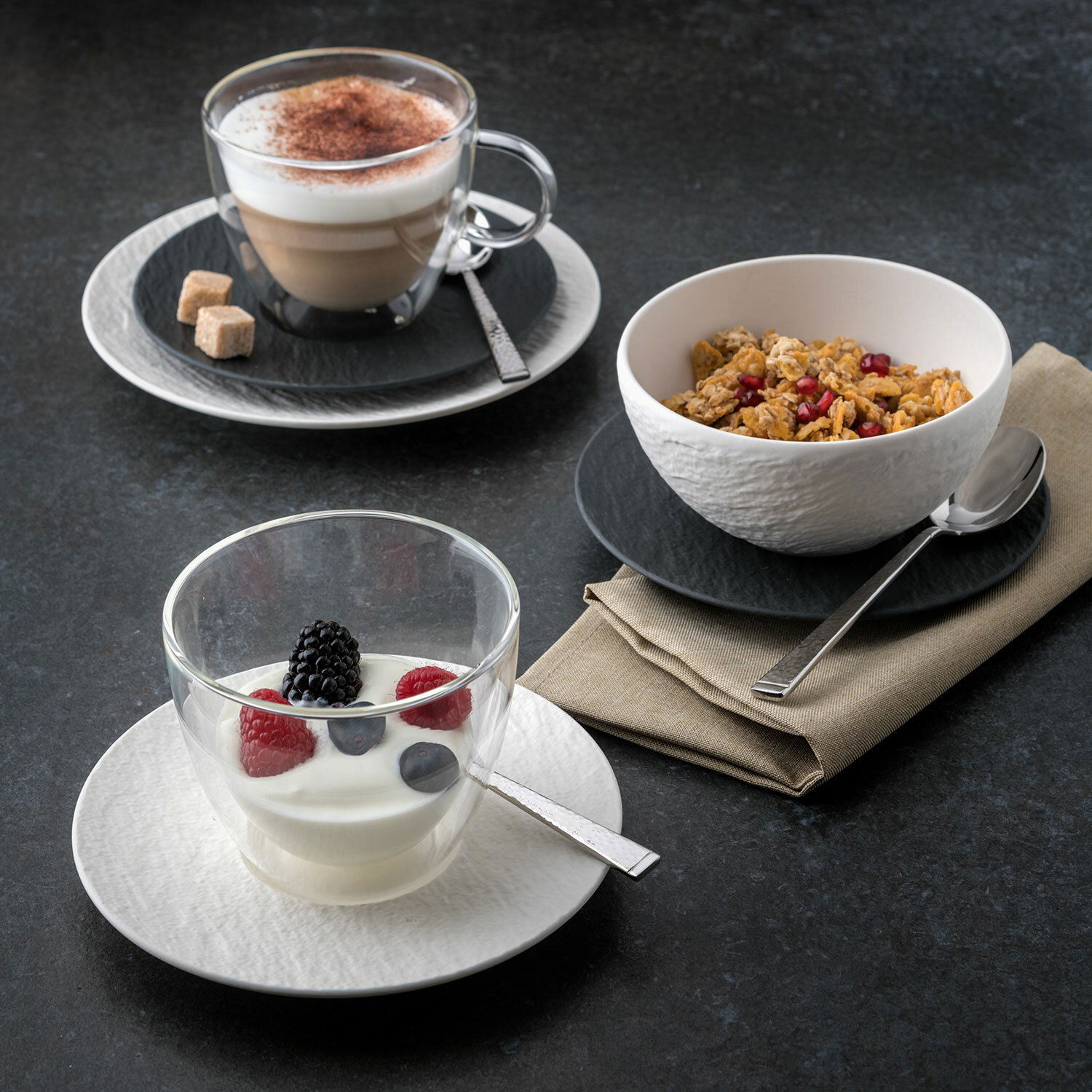 Drei stilvolle Becher auf einem dunklen Tisch, gefüllt mit Cappuccino, Joghurt mit Beeren und Müsli, aus der Vogelperspektive.