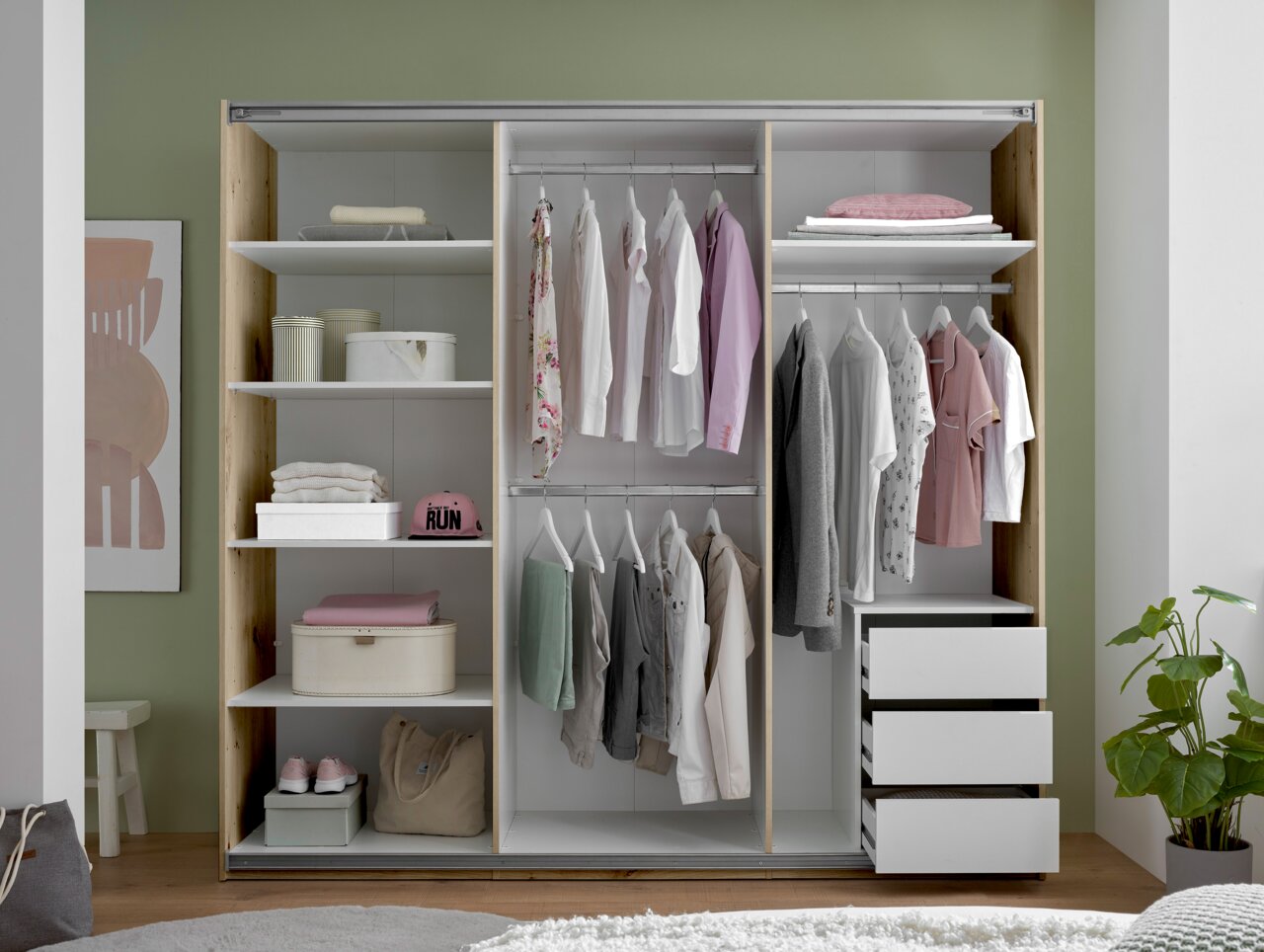 Offener Kleiderschrank mit Schiebetüren, weißer Innenausstattung, Regalböden, zwei Kleiderstangen und drei Auszügen rechts, mit Kleidung und Aufbewahrungsboxen bestückt, Frontansicht.