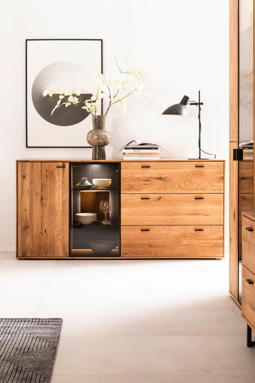 Holz-Sideboard mit Schubladen und Glastür, dekoriert mit einer Vase und Lampe, aus frontaler Perspektive.