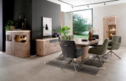 Moderne eetkamerset met een grote houten eettafel, gestoffeerde draaistoelen in groen en grijs, en vitrinekasten en een dressoir van hout met ledverlichting, gefotografeerd vanuit een schuin vooraanzicht in de lichte woonkamer.