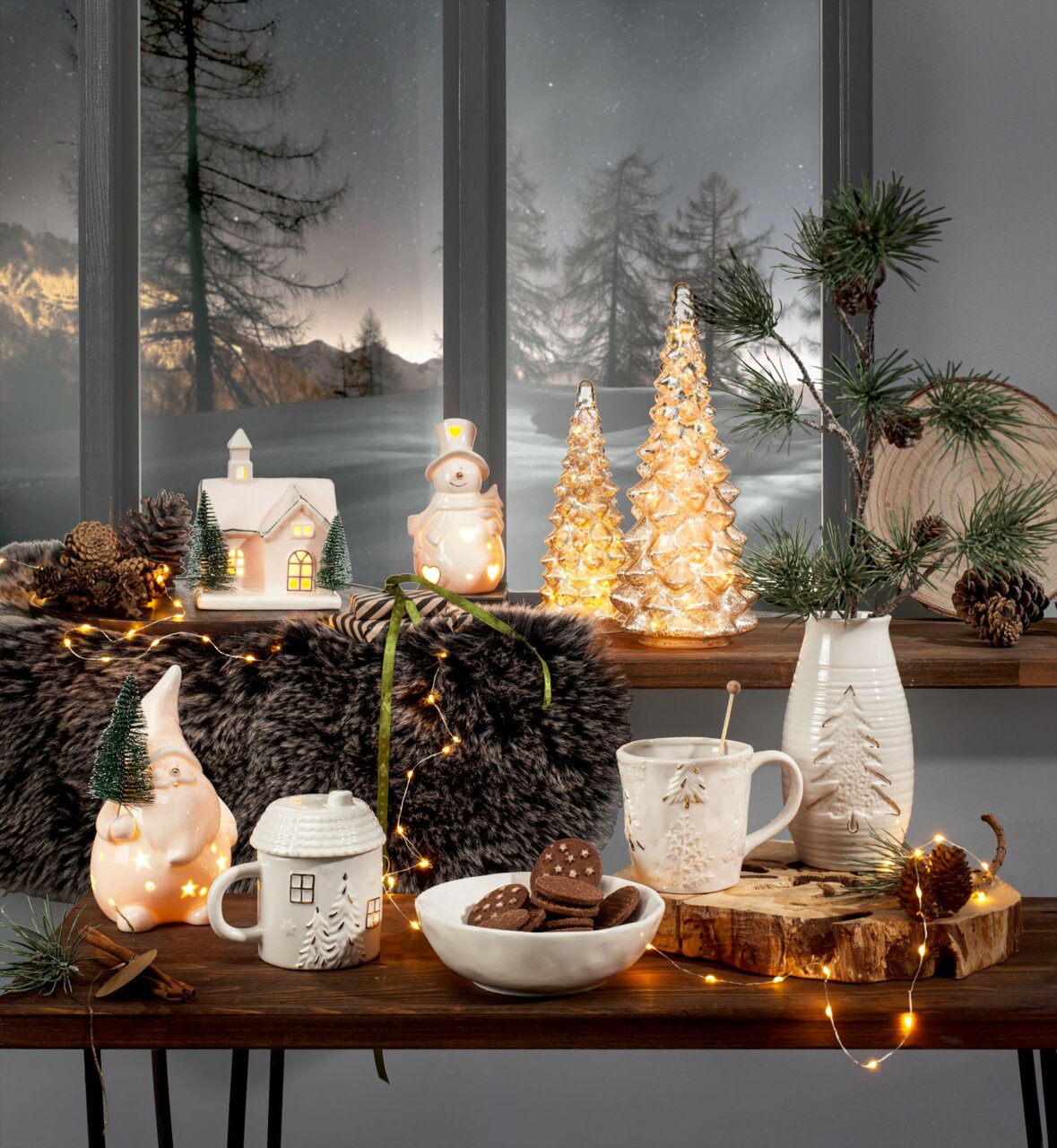 Festlich arrangierte Weihnachtsdekoration in frontaler, leicht erhöhter Perspektive mit beleuchteten Keramikfiguren, Glastannen, Tassen, Vase, Gebäck und warmweißer Lichterkette vor verschneiter Winterkulisse.
