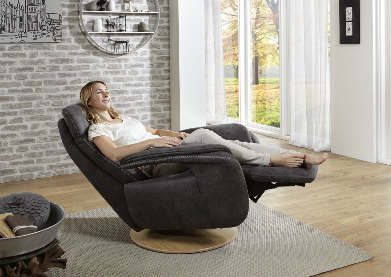 Een vrouw zit ontspannen in een moderne relaxstoel met een donkere stoffen bekleding en een houten onderstel, gezien vanuit een zijdelingse hoek, met uitzicht op een groot raam met een herfstlandschap.