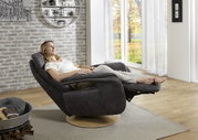 Een vrouw zit ontspannen in een moderne relaxstoel met een donkere stoffen bekleding en een houten onderstel, gezien vanuit een zijdelingse hoek, met uitzicht op een groot raam met een herfstlandschap.