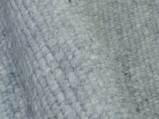 Nahaufnahme eines handgewebten Teppichs in graublauer Farbe mit strukturierter Oberfläche, seitliche Perspektive.