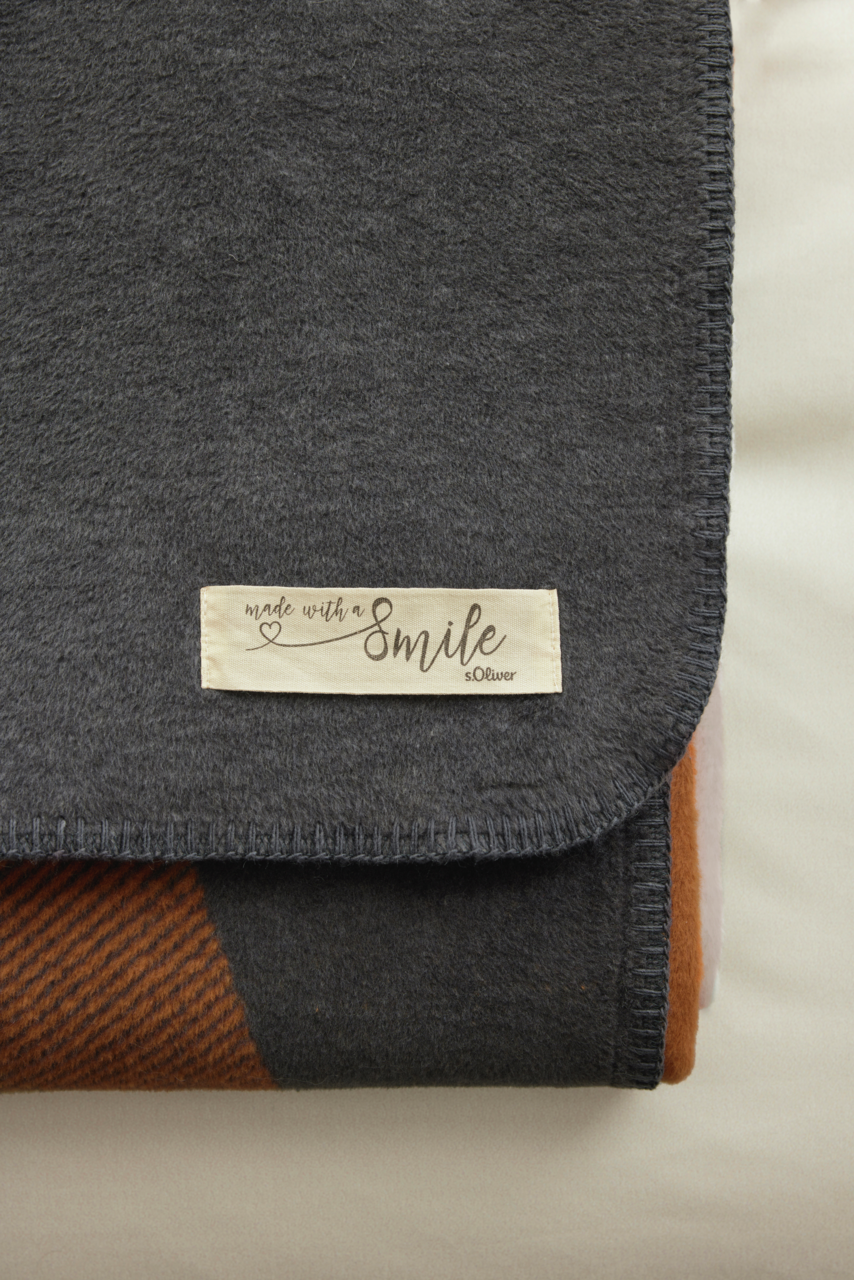 Nahaufnahme einer gefalteten Jacquard Decke von s.Oliver in dunklen Farbtönen mit einem Etikett 'made with a smile'.