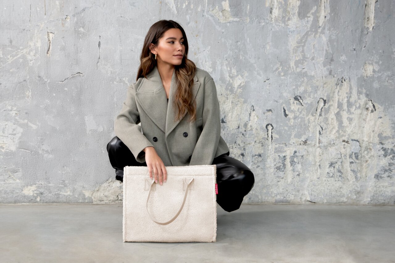 Frau in grauem Mantel und schwarzer Hose hält eine cremefarbene Tasche aus weichem Material, seitliche Perspektive