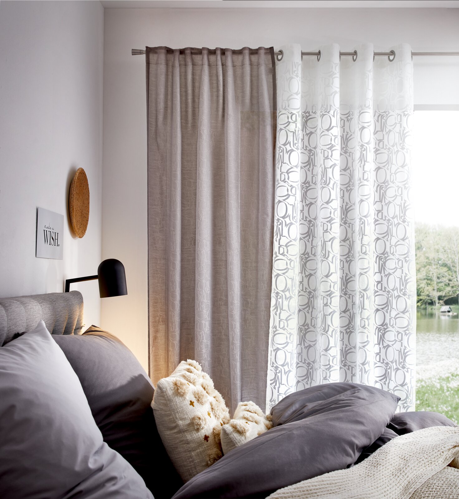 Gemütliches Schlafzimmer mit grauen und weißen Vorhängen, die durch Ösen aufgehängt sind. Die Vorhänge haben ein feines Ornamentmuster. Blick aus dem Fenster auf einen See.