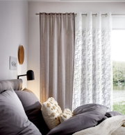 Gemütliches Schlafzimmer mit grauen und weißen Vorhängen, die durch Ösen aufgehängt sind. Die Vorhänge haben ein feines Ornamentmuster. Blick aus dem Fenster auf einen See.