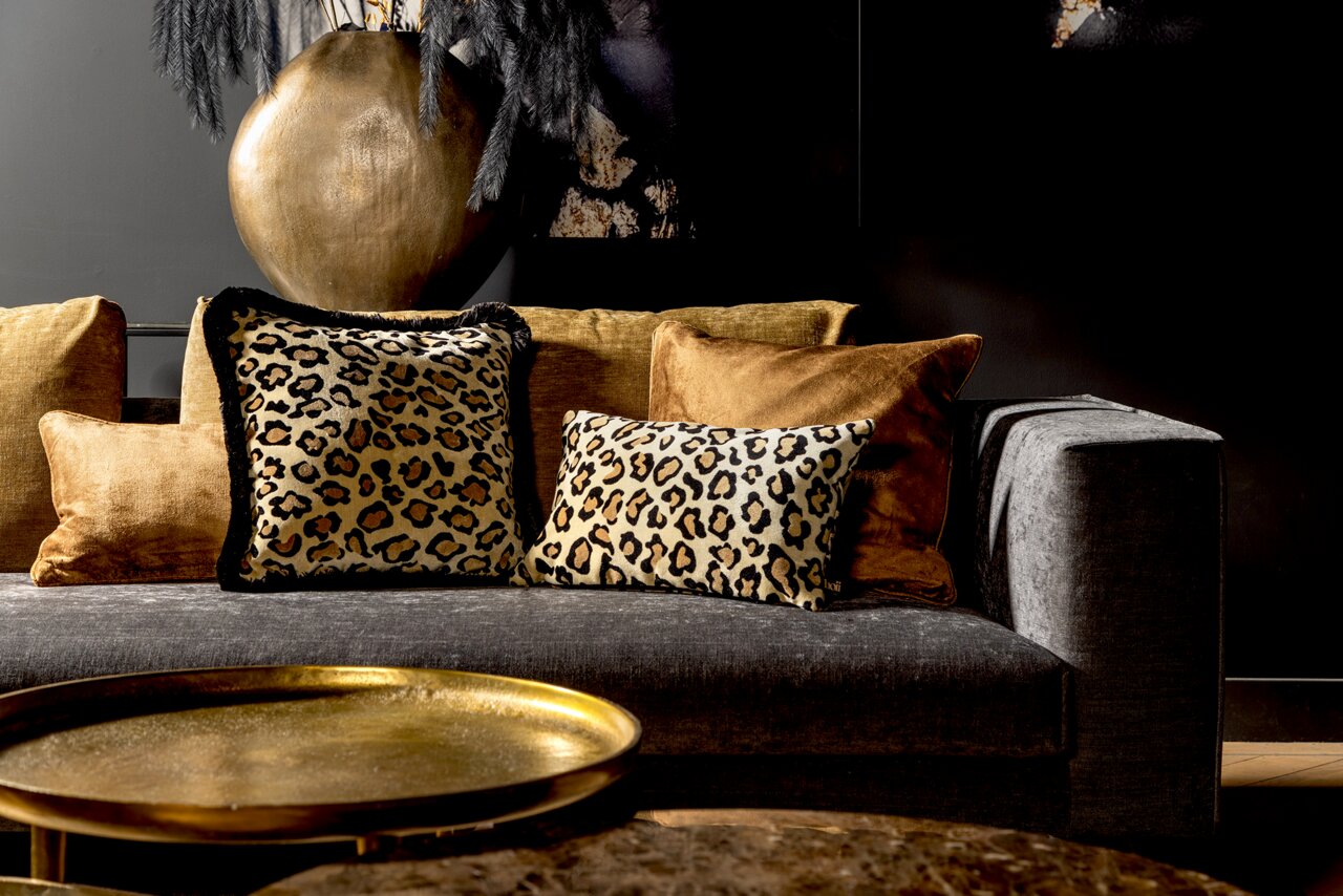 Leopardenmuster-Dekokissen auf einem grauen Sofa, umgeben von goldenen Kissen, mit einem goldenen Tisch im Vordergrund. Perspektive: Frontalansicht.