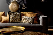Leopardenmuster-Dekokissen auf einem grauen Sofa, umgeben von goldenen Kissen, mit einem goldenen Tisch im Vordergrund. Perspektive: Frontalansicht.