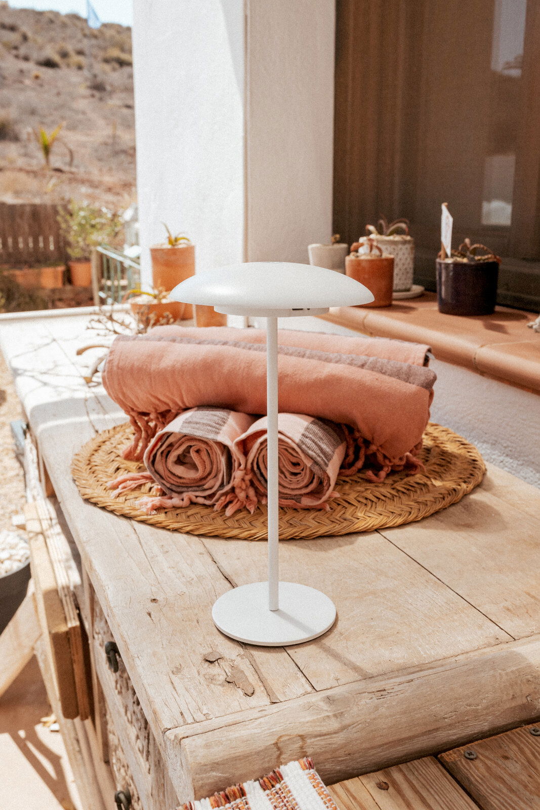 Weiße, minimalistische Tischlampe mit flachem, rundem Schirm auf rustikalem Holztisch auf einer sonnigen Terrasse, im Hintergrund gefaltete Decken und Pflanztöpfe; Perspektive: leicht von vorne und auf Augenhöhe.