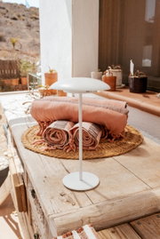 Weiße, minimalistische Tischlampe mit flachem, rundem Schirm auf rustikalem Holztisch auf einer sonnigen Terrasse, im Hintergrund gefaltete Decken und Pflanztöpfe; Perspektive: leicht von vorne und auf Augenhöhe.