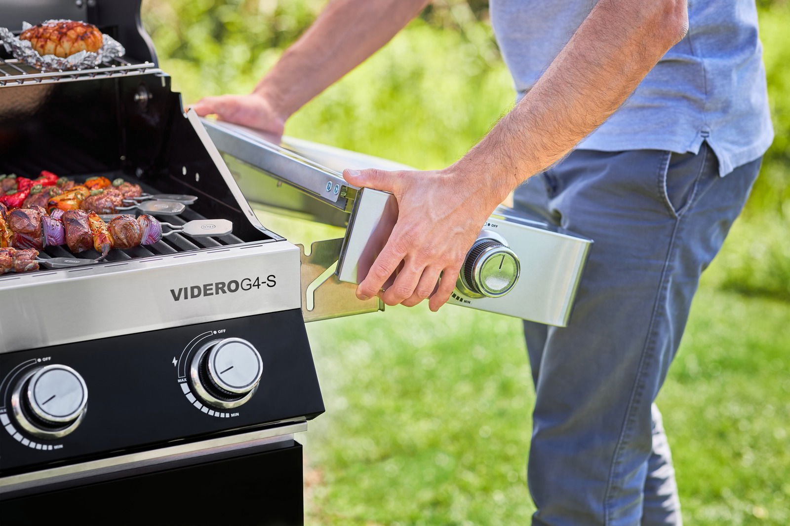 Person montiert Seitenbrenner an der BBQ-Station Videro G4-S, Vorderansicht des Grills mit gegrilltem Gemüse und Fleischspießen.