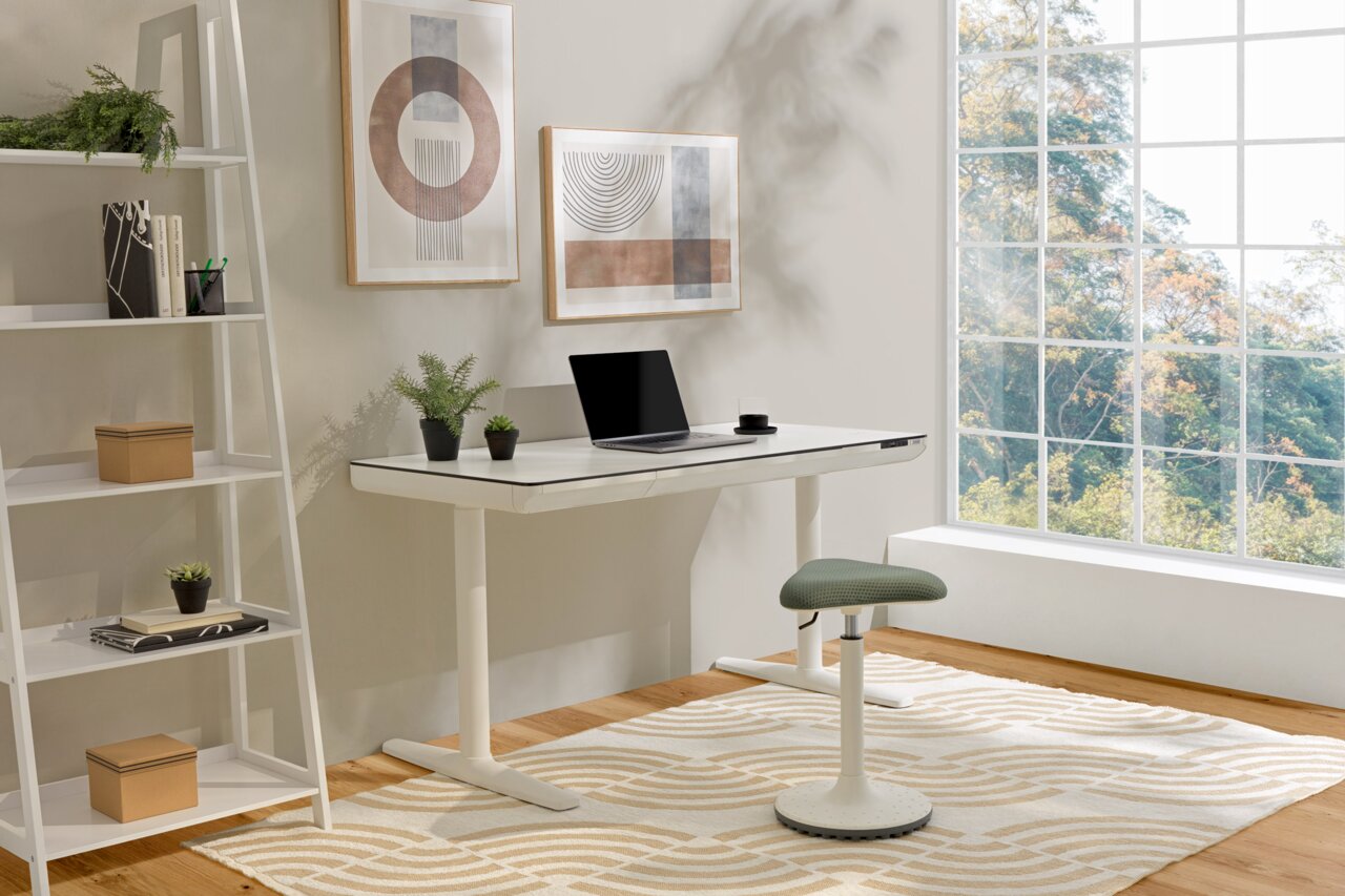 Moderner E-Schreibtisch THEA 2 in einem hellen Raum mit großem Fenster, seitliche Perspektive. Der Schreibtisch ist weiß mit einem Laptop und Pflanzen darauf, daneben ein ergonomischer Hocker.