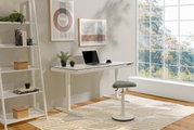 Moderner E-Schreibtisch THEA 2 in einem hellen Raum mit großem Fenster, seitliche Perspektive. Der Schreibtisch ist weiß mit einem Laptop und Pflanzen darauf, daneben ein ergonomischer Hocker.