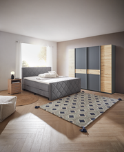 Gemütliches Schlafzimmer mit Boxspringbett BX2200 CHESTER in grauem Stoff, frontal aufgenommen. Neben dem Bett steht ein Nachttisch mit Lampe, im Hintergrund ein Kleiderschrank mit Holz- und Grauelementen.