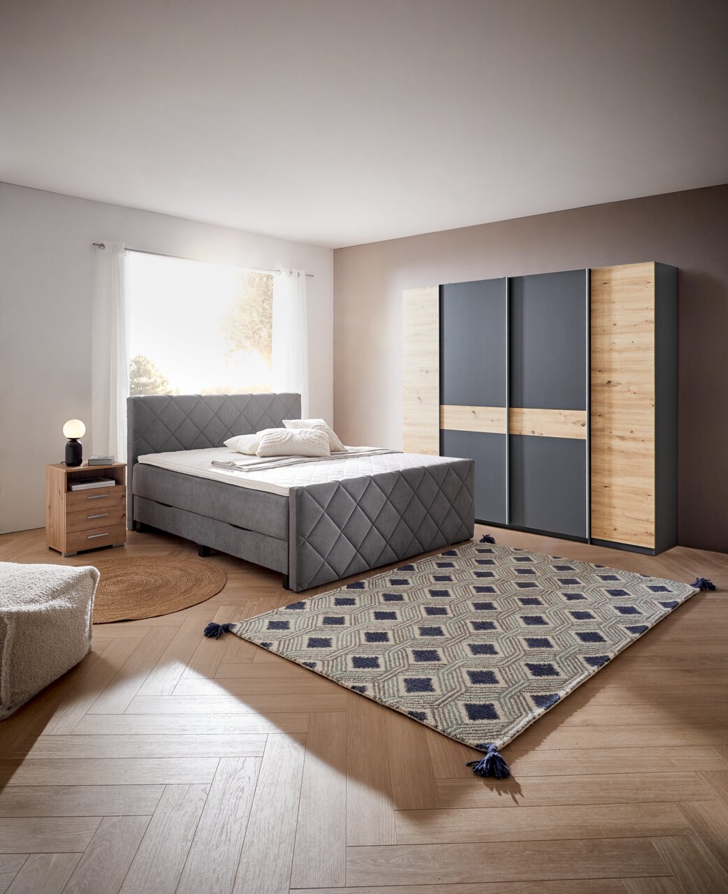 Gemütliches Schlafzimmer mit Boxspringbett BX2200 CHESTER in grauem Stoff, frontal aufgenommen. Neben dem Bett steht ein Nachttisch mit Lampe, im Hintergrund ein Kleiderschrank mit Holz- und Grauelementen.