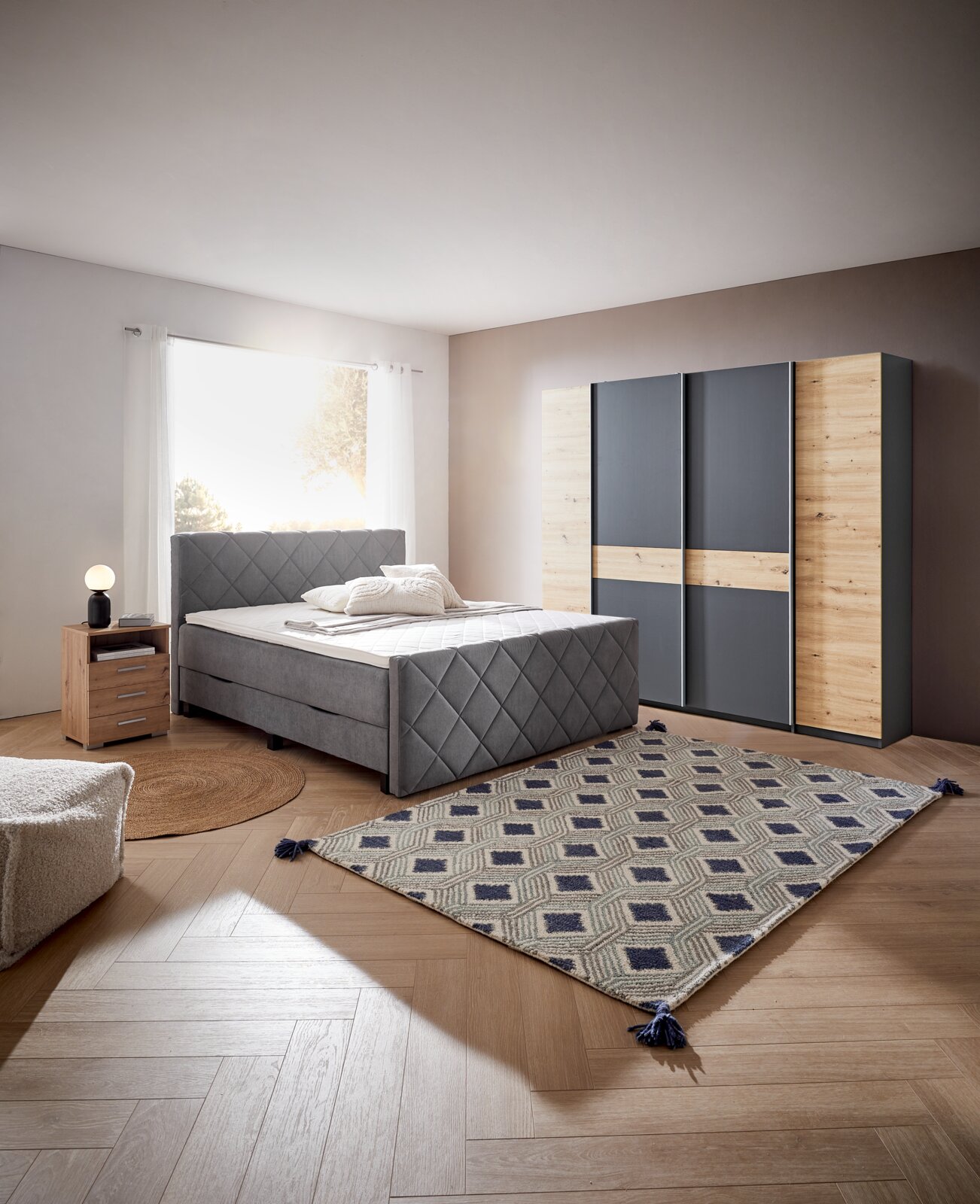 Gezellige slaapkamer met boxspring BX2200 CHESTER in grijze stof, gefotografeerd vanaf de voorkant. Naast het bed staat een nachtkastje met lamp, op de achtergrond een kledingkast met houten en grijze elementen.