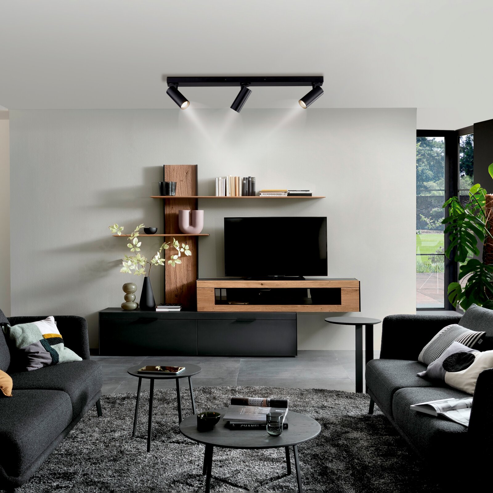 Modernes Wohnzimmer mit schwarzer Decken-Spotleuchte mit drei Strahlern über TV-Board und Wandregal, frontale Perspektive mit leichtem Blick nach oben