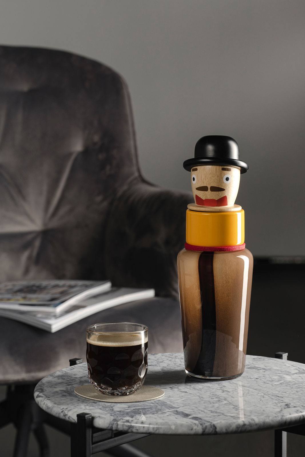Leonardo Luigi-vaas BUONI AMICI Objektvase Luigi in Form einer stilisierten Figur mit Hut, auf einem Marmortisch neben einem Glas Kaffee, im Hintergrund ein Sessel. Perspektive: seitlich.