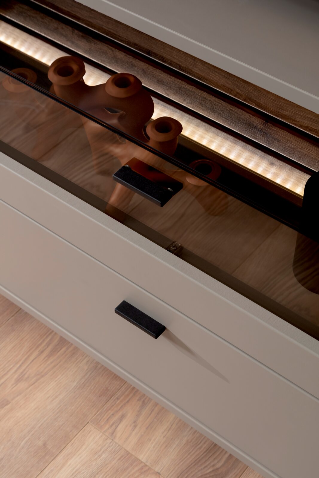 Zurbrüggen Tv-element LÜTTICH Een detailfoto, schuin van bovenaf genomen, van een modern dressoir met een grijze voorkant, een zwarte handgreep en een vitrine van getint glas, waarin een terracottakleurige keramische vaas staat.