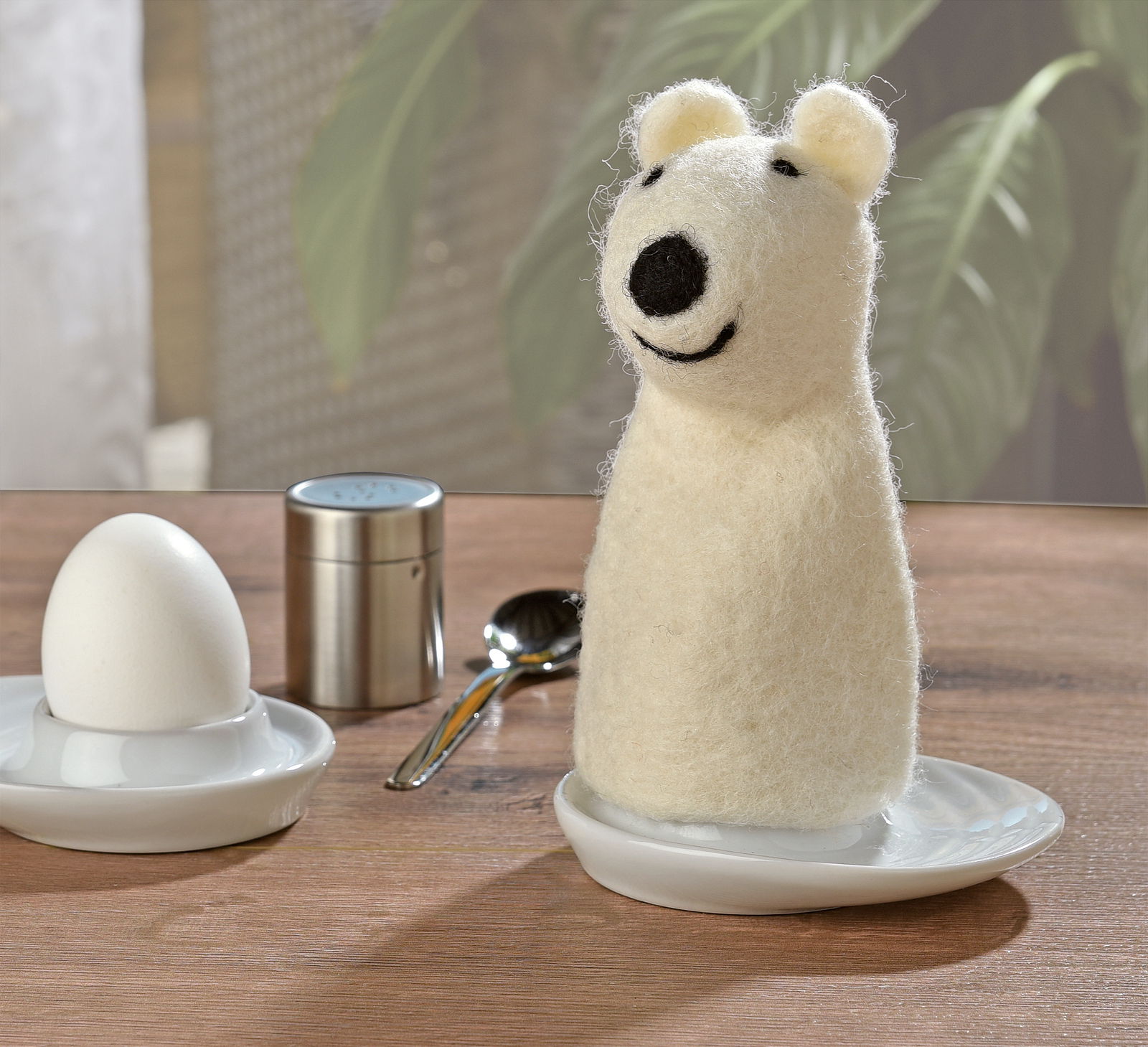 Eierwärmer in Form eines lächelnden Eisbären aus Filz, stehend auf einem Tisch neben einem Ei und einem Löffel, seitliche Perspektive.