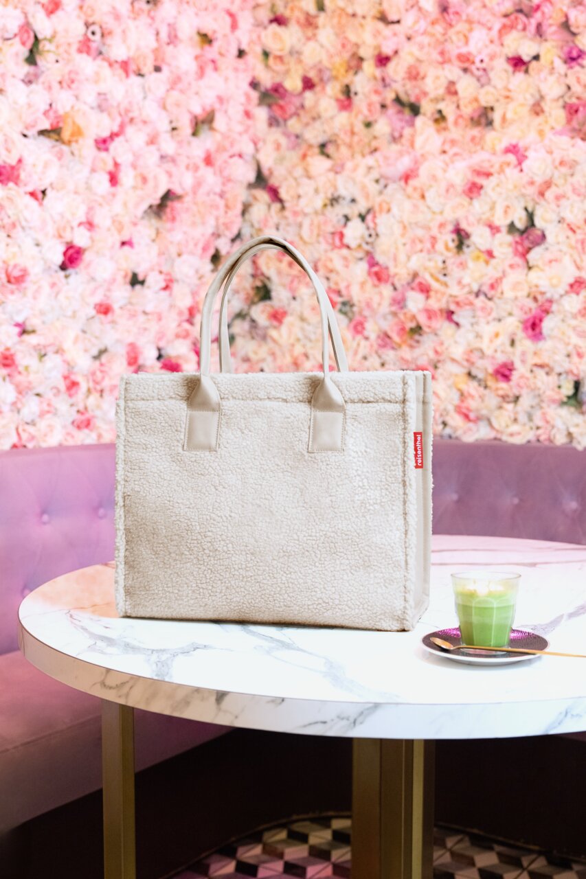 Beige shopper van zacht materiaal op een marmeren tafel, omringd door een bloemrijke wanddecoratie, gezien van voren.