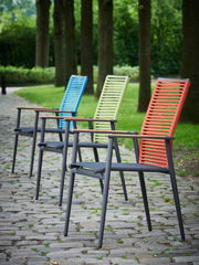 Vier bunte Gartenstühle in einer Reihe auf einem gepflasterten Weg, umgeben von Bäumen, aus seitlicher Perspektive fotografiert.