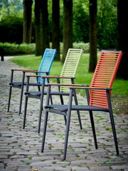 Vier bunte Gartenstühle in einer Reihe auf einem gepflasterten Weg, umgeben von Bäumen, aus seitlicher Perspektive fotografiert.
