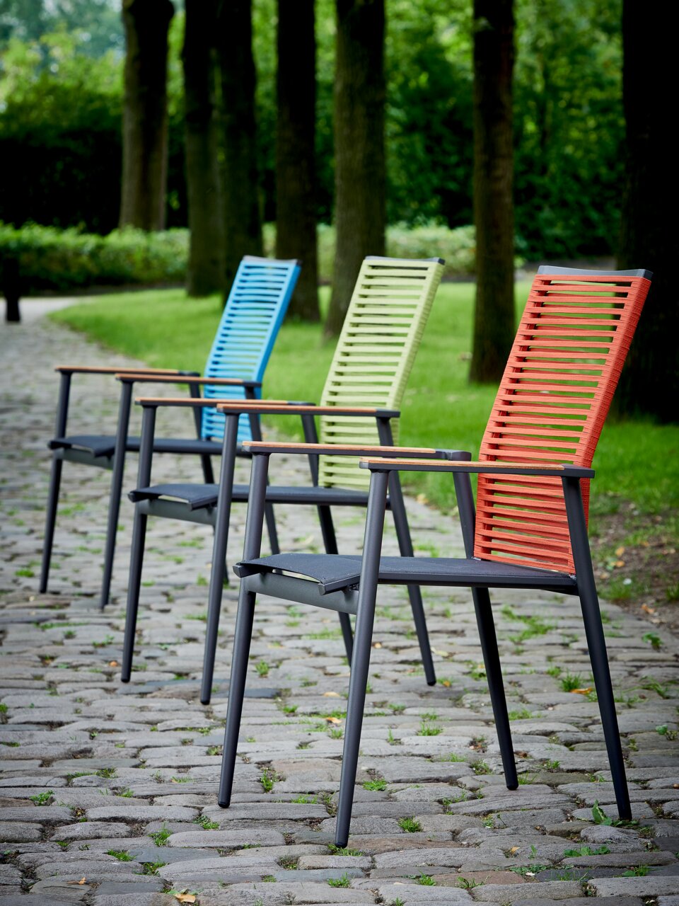 Vier kleurrijke tuinstoelen staan op een rij op een geplaveid pad, omringd door bomen, gefotografeerd vanuit een zijdelingse hoek.