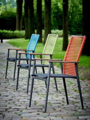 Vier kleurrijke tuinstoelen staan op een rij op een geplaveid pad, omringd door bomen, gefotografeerd vanuit een zijdelingse hoek.