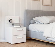 Weißes Nachttisch-Paneel mit zwei Schubladen, einem Regal und einer Steckdose, seitliche Perspektive neben einem grauen Bett.