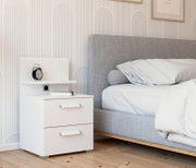 Weißes Nachttisch-Paneel mit zwei Schubladen, einem Regal und einer Steckdose, seitliche Perspektive neben einem grauen Bett.