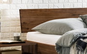 Close-up van een houten bedframe met hoofdeinde, grijs beddengoed en een nachtkastje met een kopje en een boek.