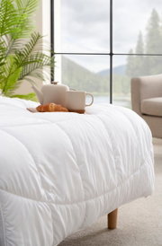 Weiße Luxe Leicht-Decke auf einem Bett, mit einer Teekanne, Tasse und Croissant darauf, im Vordergrund. Im Hintergrund ein großes Fenster mit Blick auf eine grüne Landschaft.