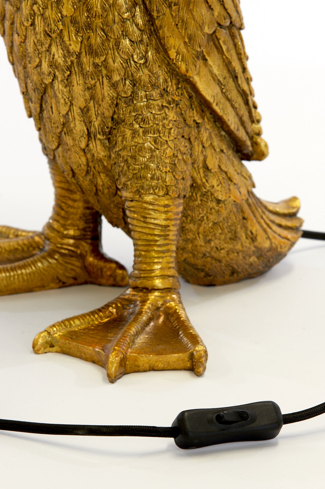 Close-up van de gouden voet van een tafellamp in de vorm van een vogel, met een zwarte kabel en een schakelaar op de voorgrond.