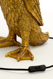 Close-up van de gouden voet van een tafellamp in de vorm van een vogel, met een zwarte kabel en een schakelaar op de voorgrond.