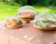 Drei runde Abdeckhauben aus Bambus mit Netz, die auf einem Tisch im Freien stehen. Sie schützen Obst wie Orangen, Melonen und Trauben vor Insekten. Die Perspektive zeigt die Abdeckhauben von schräg oben.