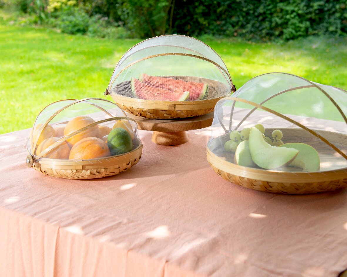 Decoris Vliegenkapje, set van 3 Drie ronde afdekkappen van bamboe met gaas, die op een tafel buiten staan. Ze beschermen fruit zoals sinaasappels, meloenen en druiven tegen insecten. Het perspectief toont de afdekkappen vanuit een schuine hoek van bovenaf.